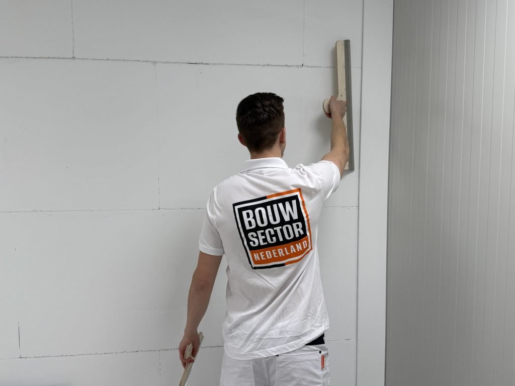 Achteraanzicht van een stukadoor met logo die een rei verticaal tegen de wand houdt