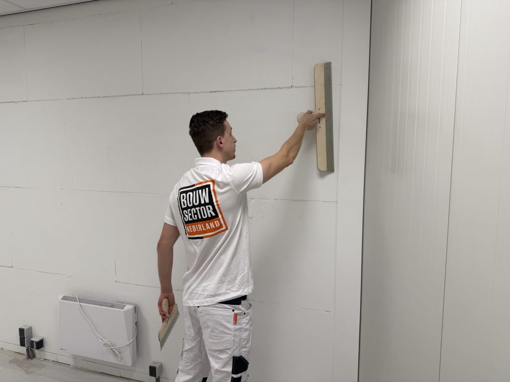 Stukadoor strekt zijn arm om het bovenste deel van een wand vlak te trekken, naast een kachel op de vloer