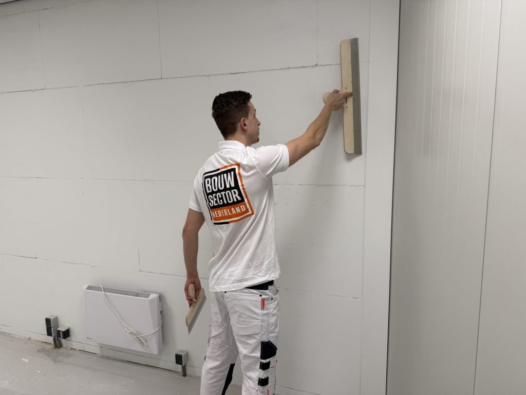 Vakman in wit werkpak zet een stucrei hoog tegen de muur bij de hoek van de ruimte