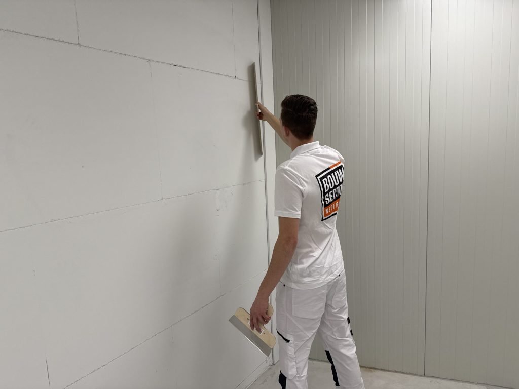 Stukadoor werkt langs een lange wand van grote blokken en houdt een spaan klaar in zijn linkerhand