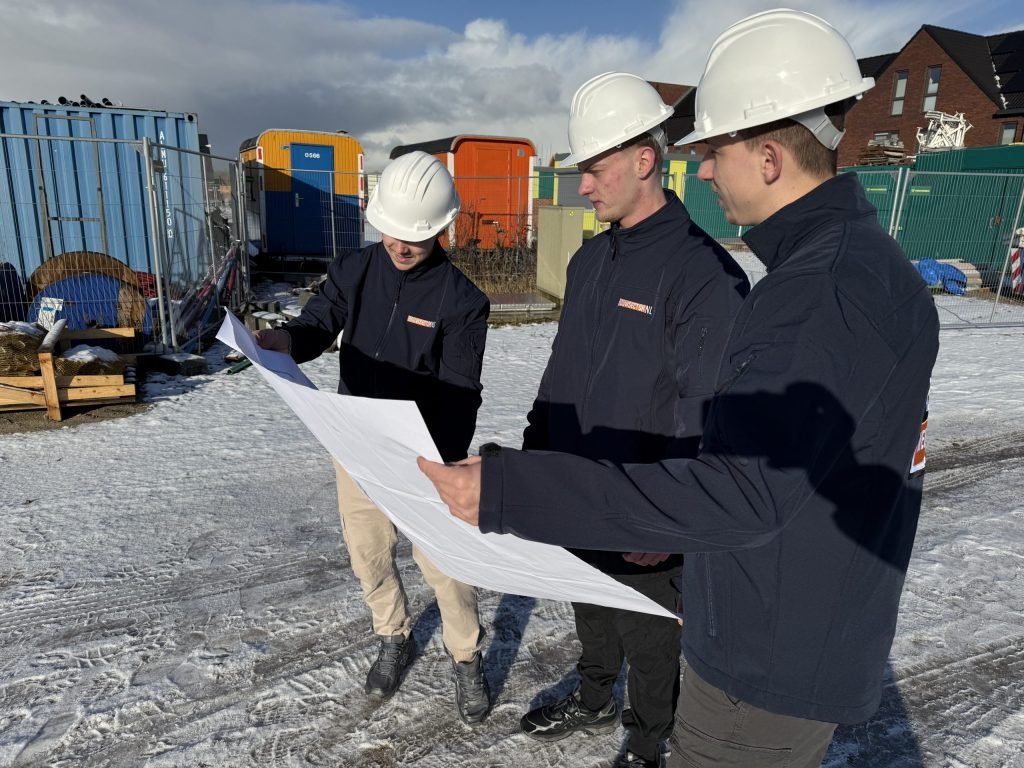 Drie vakmensen van Bouwsector Nederland met witte helm bekijken samen een bouwtekening op een nieuwbouwterrein